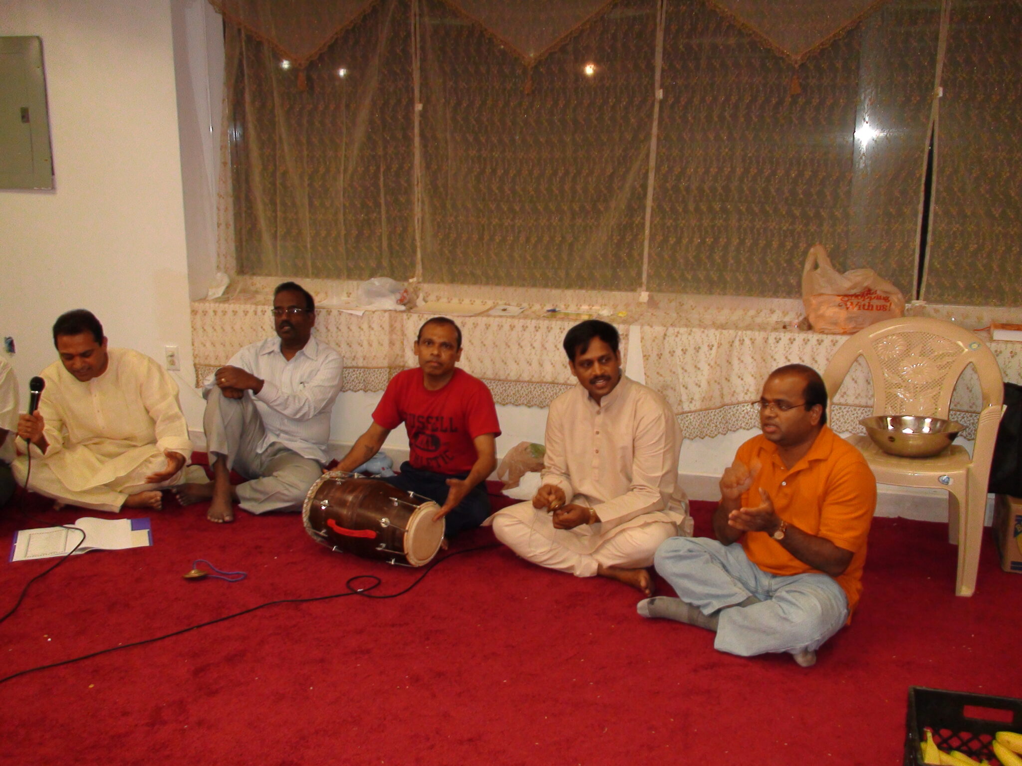 Five men seated on a red carpet performing music and devotional songs at a Ganesh Chaturthi gathering